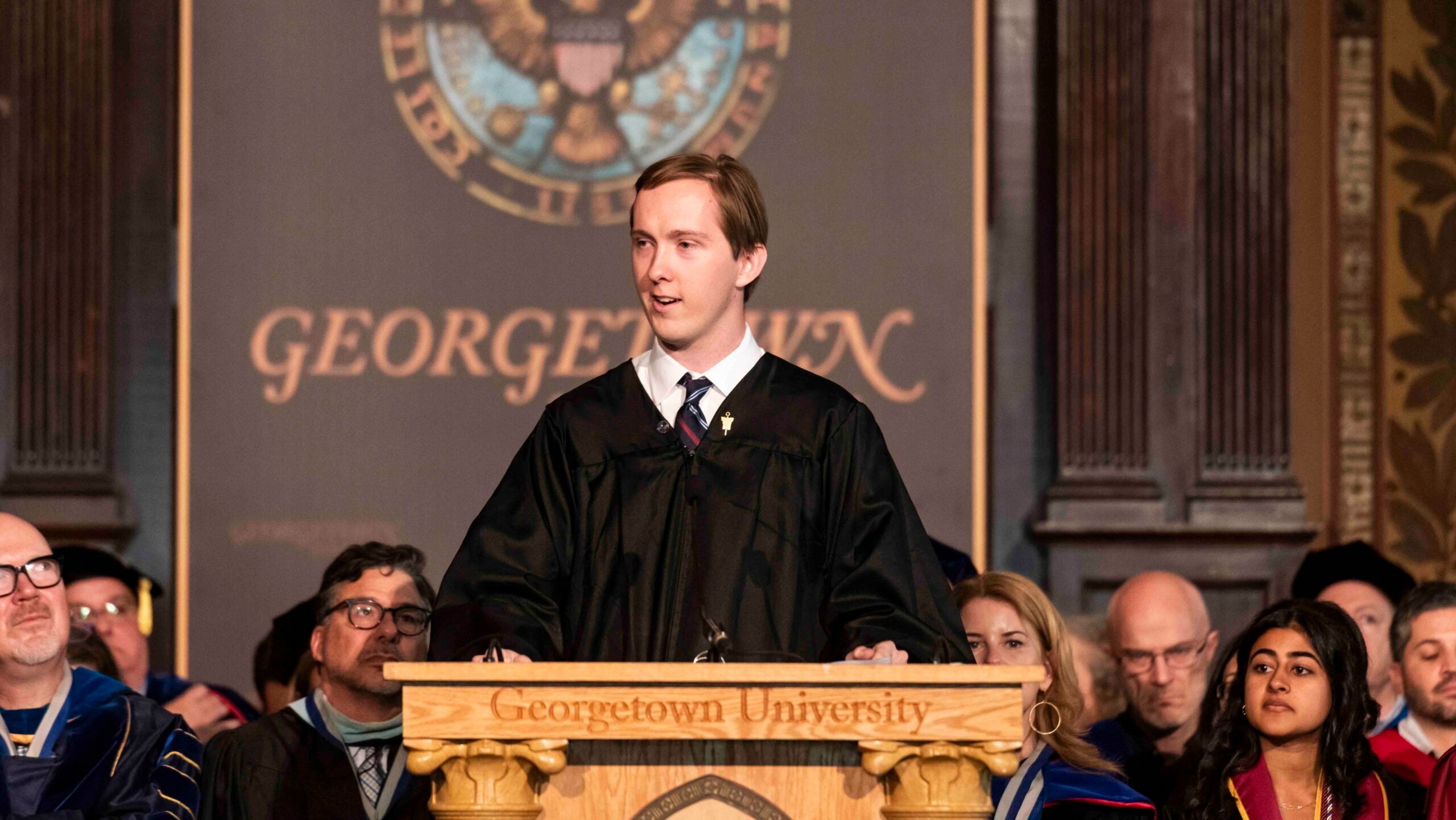 A young man in academic regalia speaks at a podium. He wears a shirt with a white collar and a tie. His hair is combed.