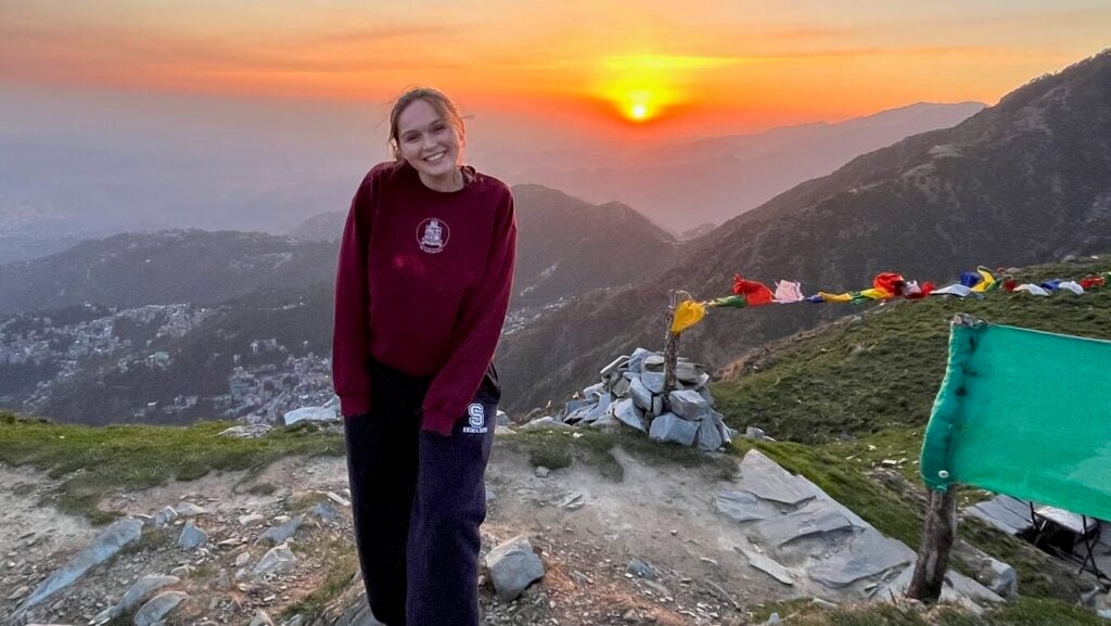 A girl with her hair back smiles at the camera. She wears loose sweat pants and a sweatshirt and stands on the top of a mountain. Behind her, the sun rises over a wide-reaching valley.