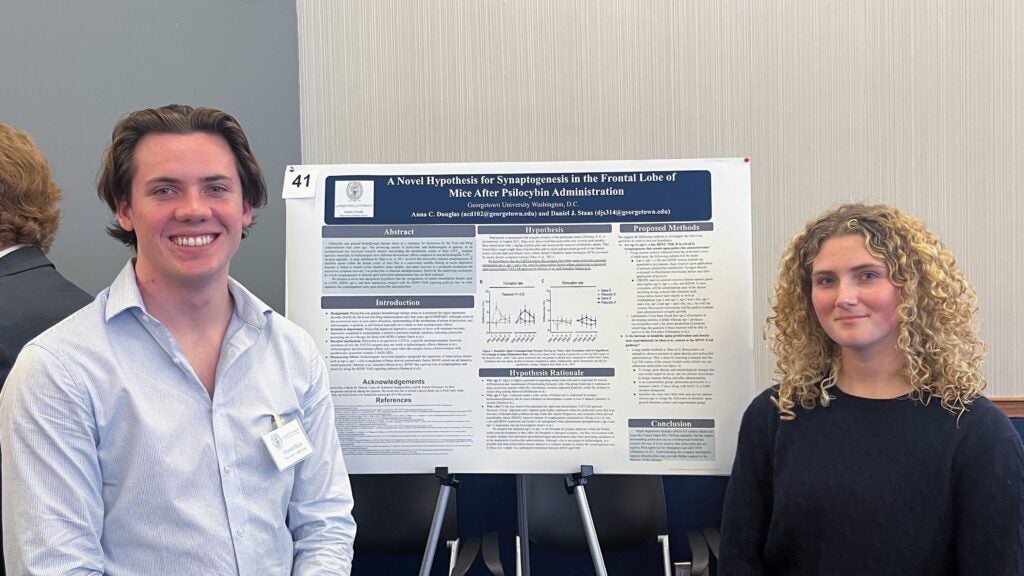 Two people stand next to a poster board at a research conference. One, on the right, has curly, blonde hair. The other, on the left, has short hair and wears a button-down shirt.