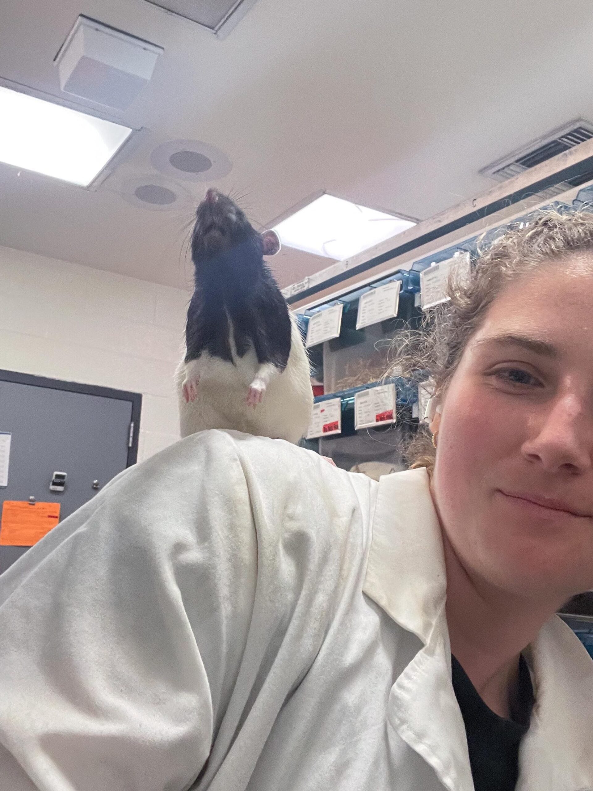 A girl in a white lab coat takes a selfie with a rat on her shoulder.