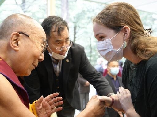 On the left is the Dalai Lama, a bad man wearing an orange robe and eye glasses. He shakes hands with a woman in a white dress wearing a white mask.