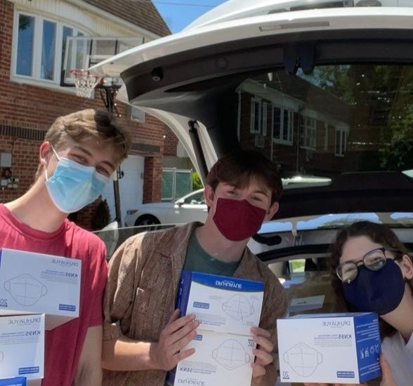 Three young people stand in front of an open hatchback car. They are wearing masks and holding boxes of masks in their hands.