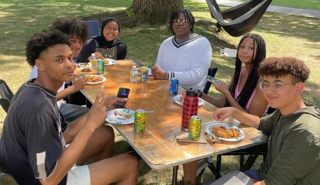A group of college students sit around a picnic table outside and share a meal. 