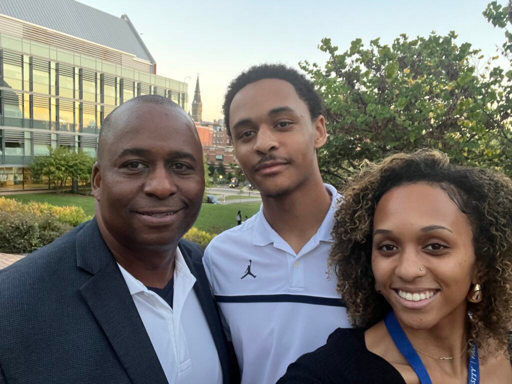 A family of three smiles and takes a selfie outside. The father wears professional attire and the daughter and son wear casual clothes.