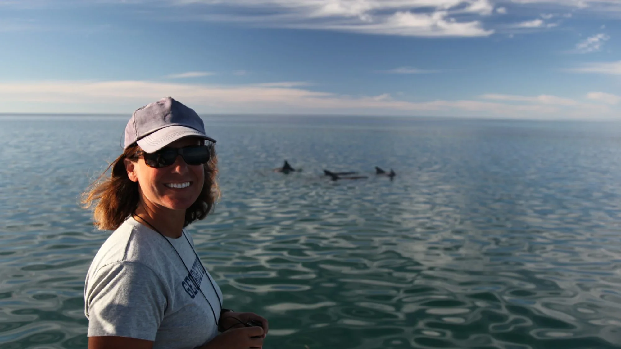A woman with medium-length dark hair wears a gray Georgetown tee shirt and a gray hat. She is also wearing sunglasses and smiling at the camera. Behind her is a wide body of water.