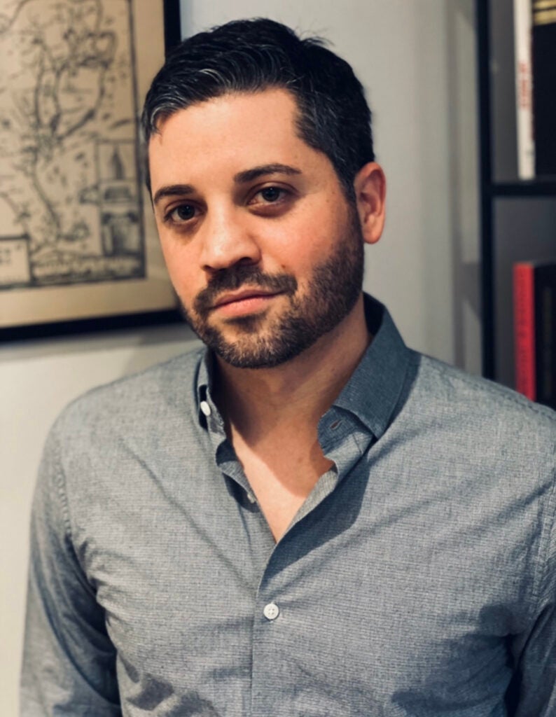 A professor in a buttoned shirt looks directly at the camera.