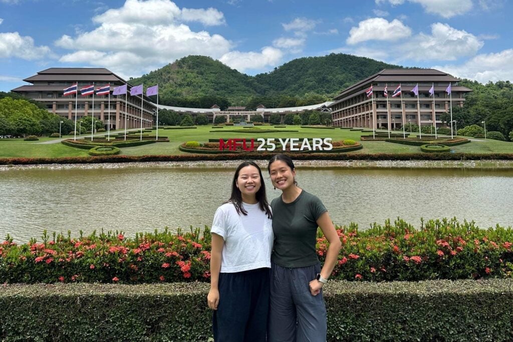 Two girls stand in front of a lake with a large college building on the opposite shore befind them.