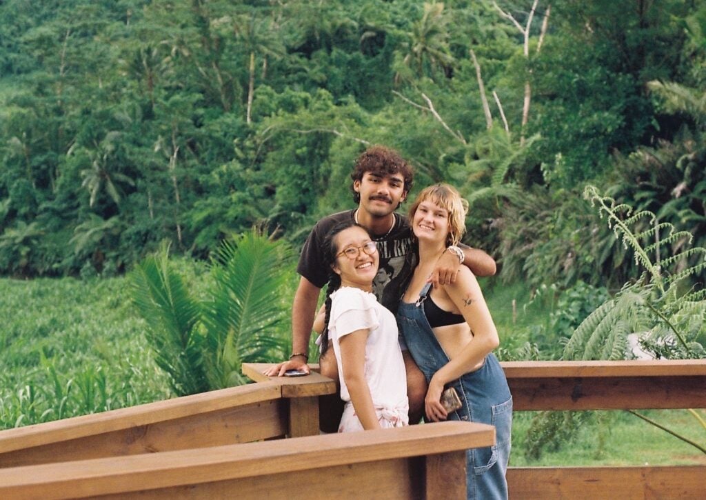 Three young people stand on a wooden board walk in front of a verdant, green forest.