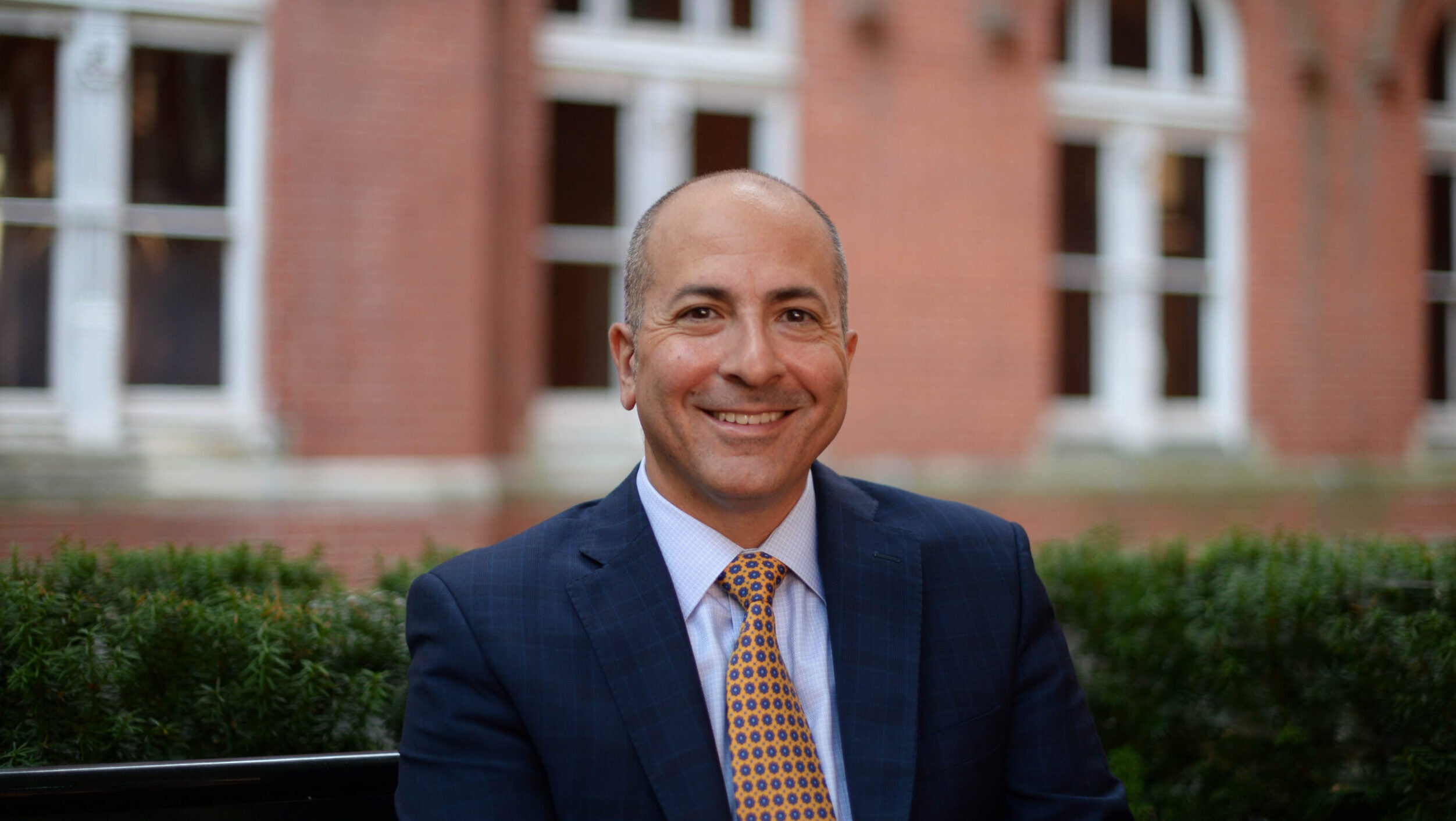 A man sits outside and smiles facing the camera. Behind him is an out-of-focus red brick wall. He wears a white button down shirt, a navy jacket, and a red tie.