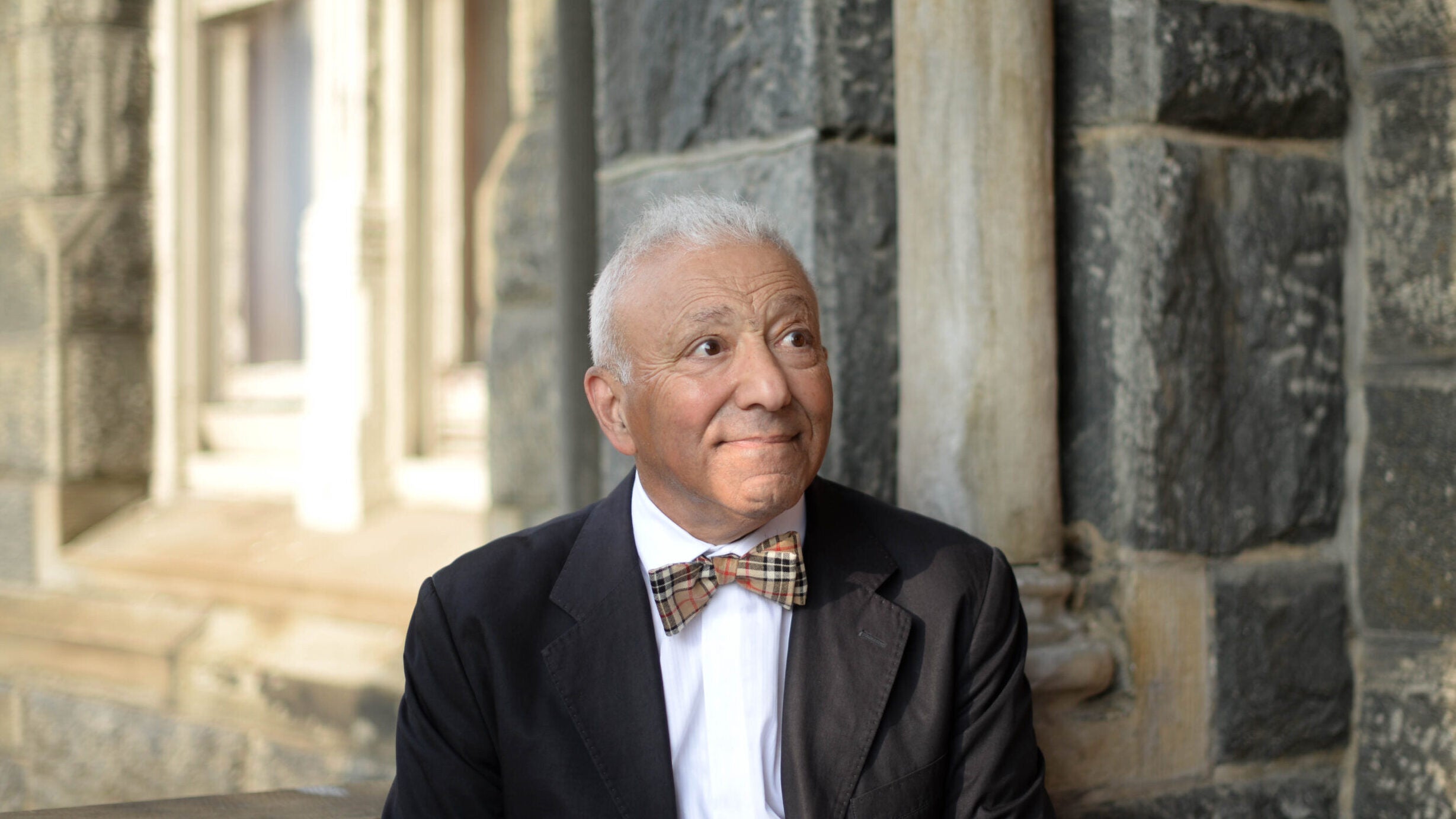 A man in a black jacket, white shirt, and colorful bow tie softly smiles. He looks off camera, to his left, and stands in front of an out-of-focus gray stone building.