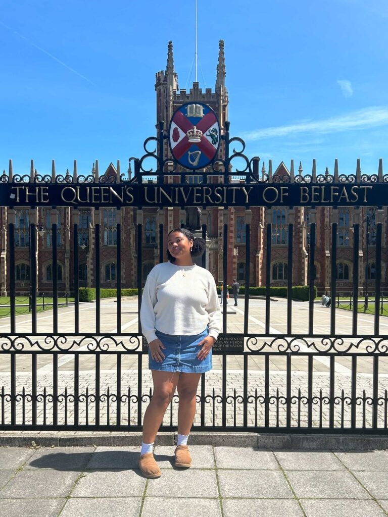 A girl with a white top and blue skirts stands in front of wrought iron gates that read 'Queen's University Belfast.' Behind the gates are a gray stone building.