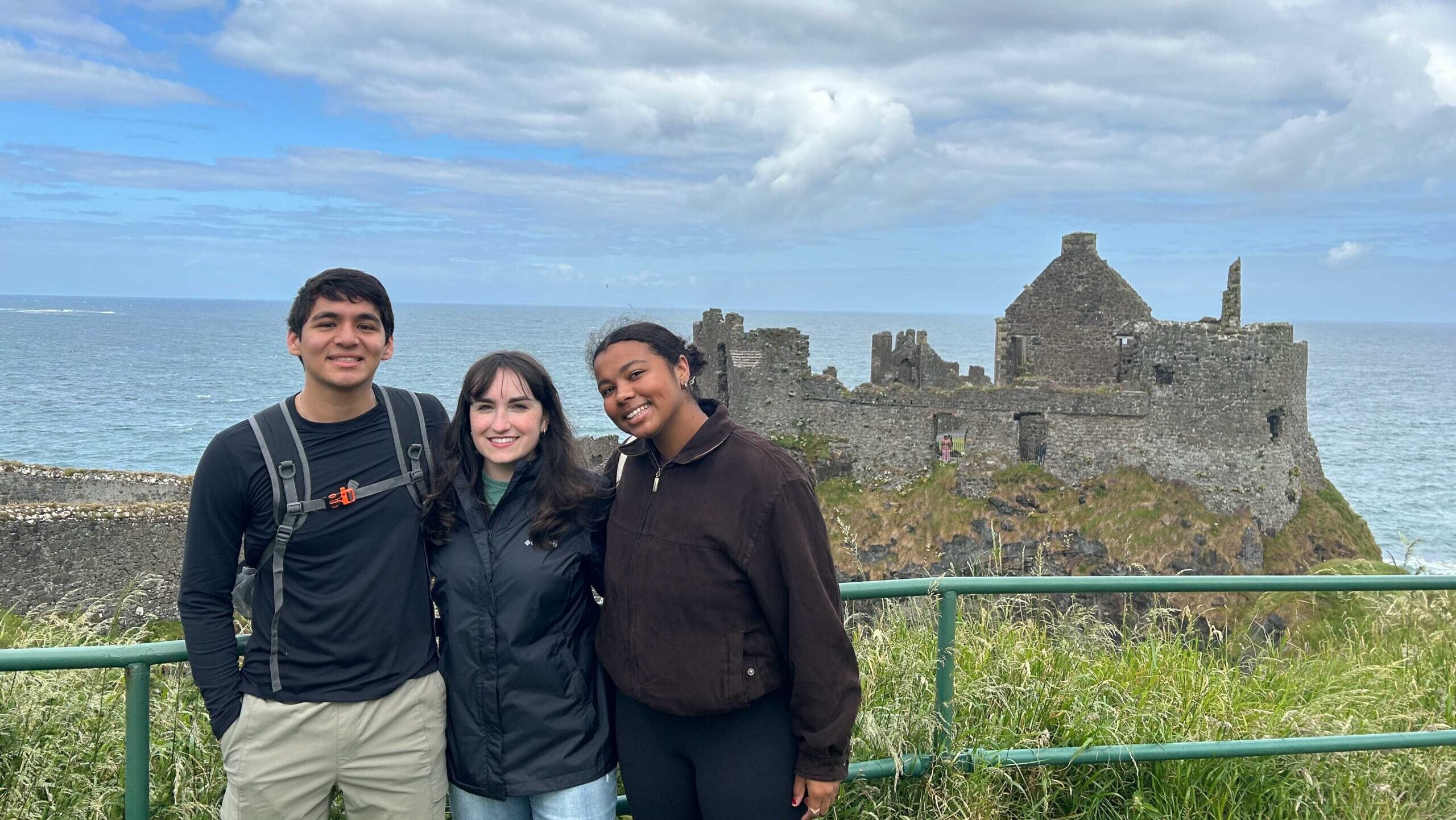 Three young adults stand in front of the ruins of a shorefront castle.