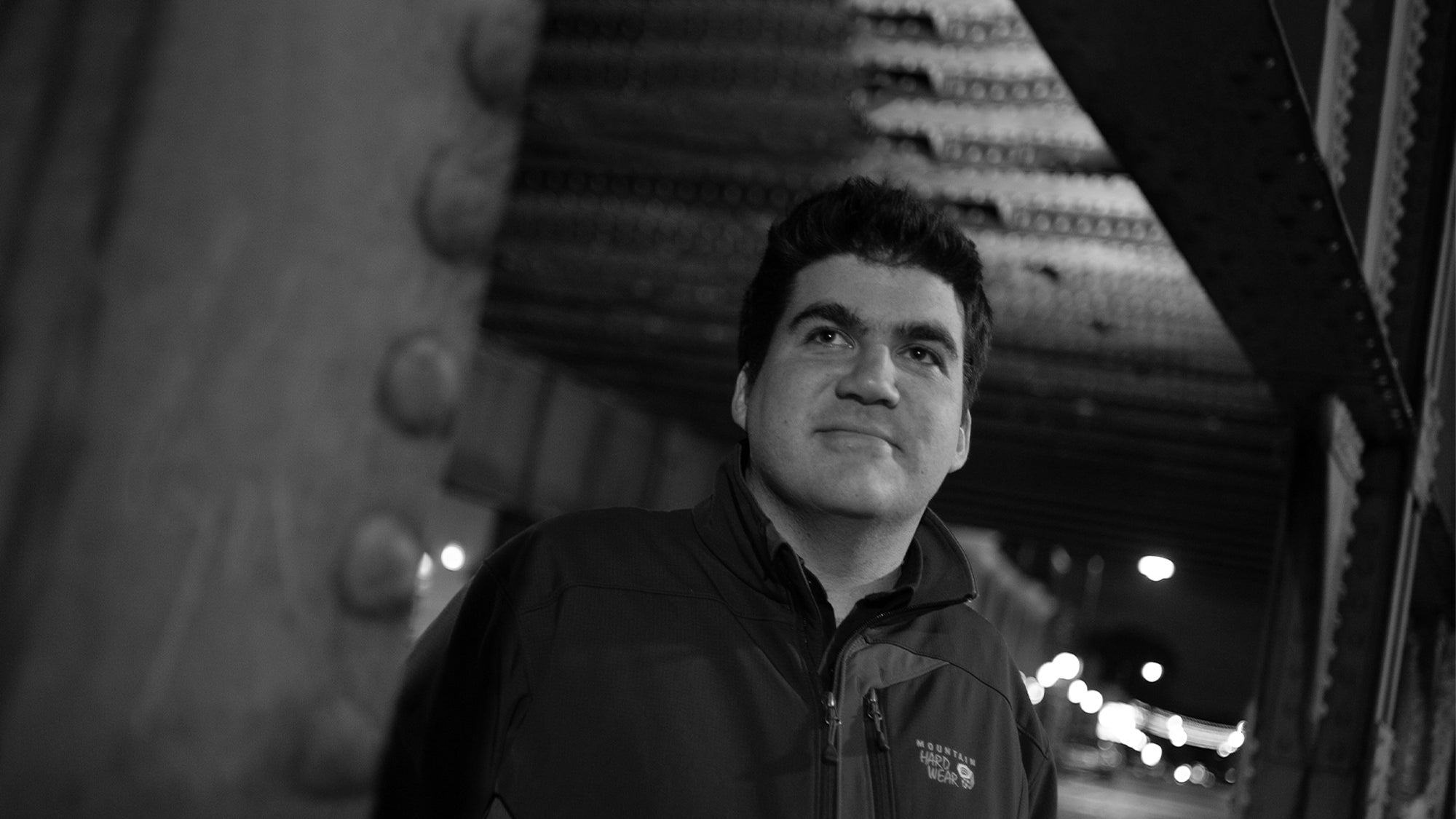 A black and white image of a man in a hooded jacket standing underneath an overpass.