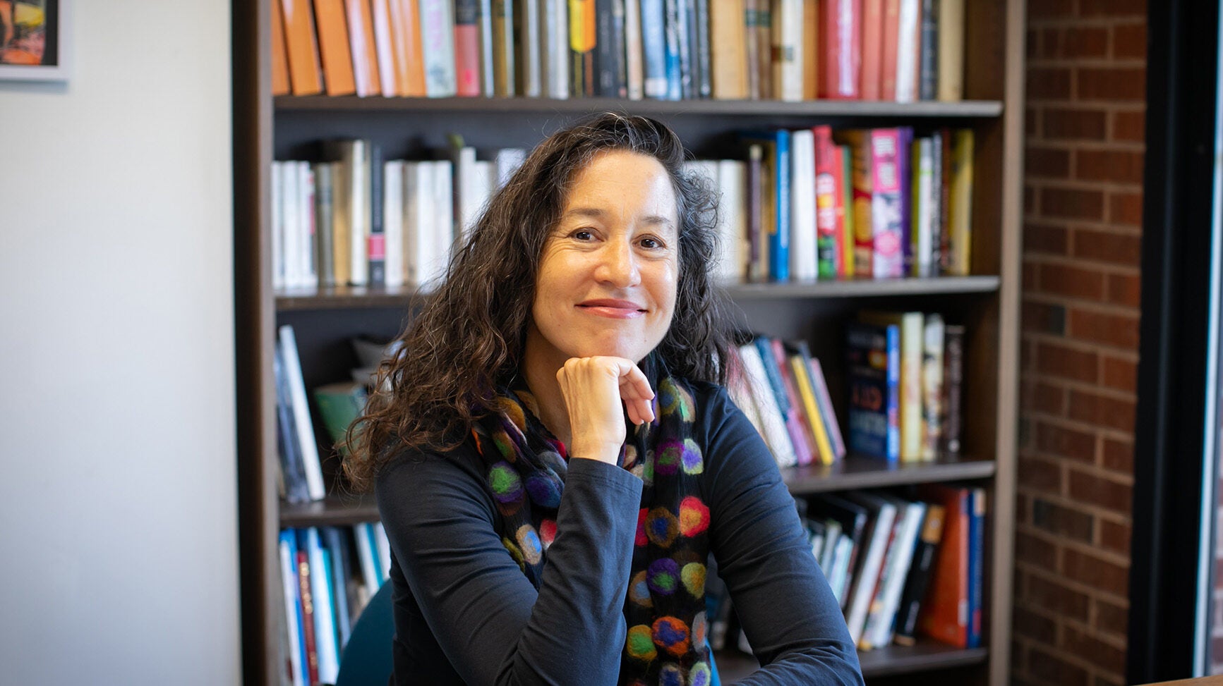 A woman with long, black, curly hair sits at her desk and smiles. She has a fist under her chin and her glasses sit on the table by her side. Behind her is a large, out-of-focus book shelf.