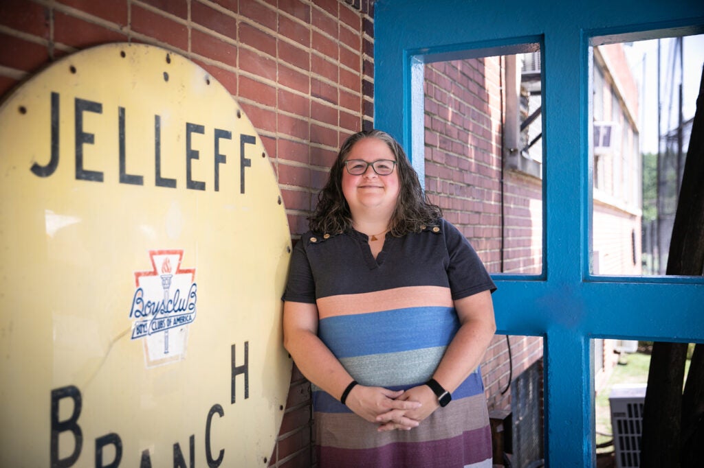 A bespectacled woman with a striped shirt stands in front of a brick wall.