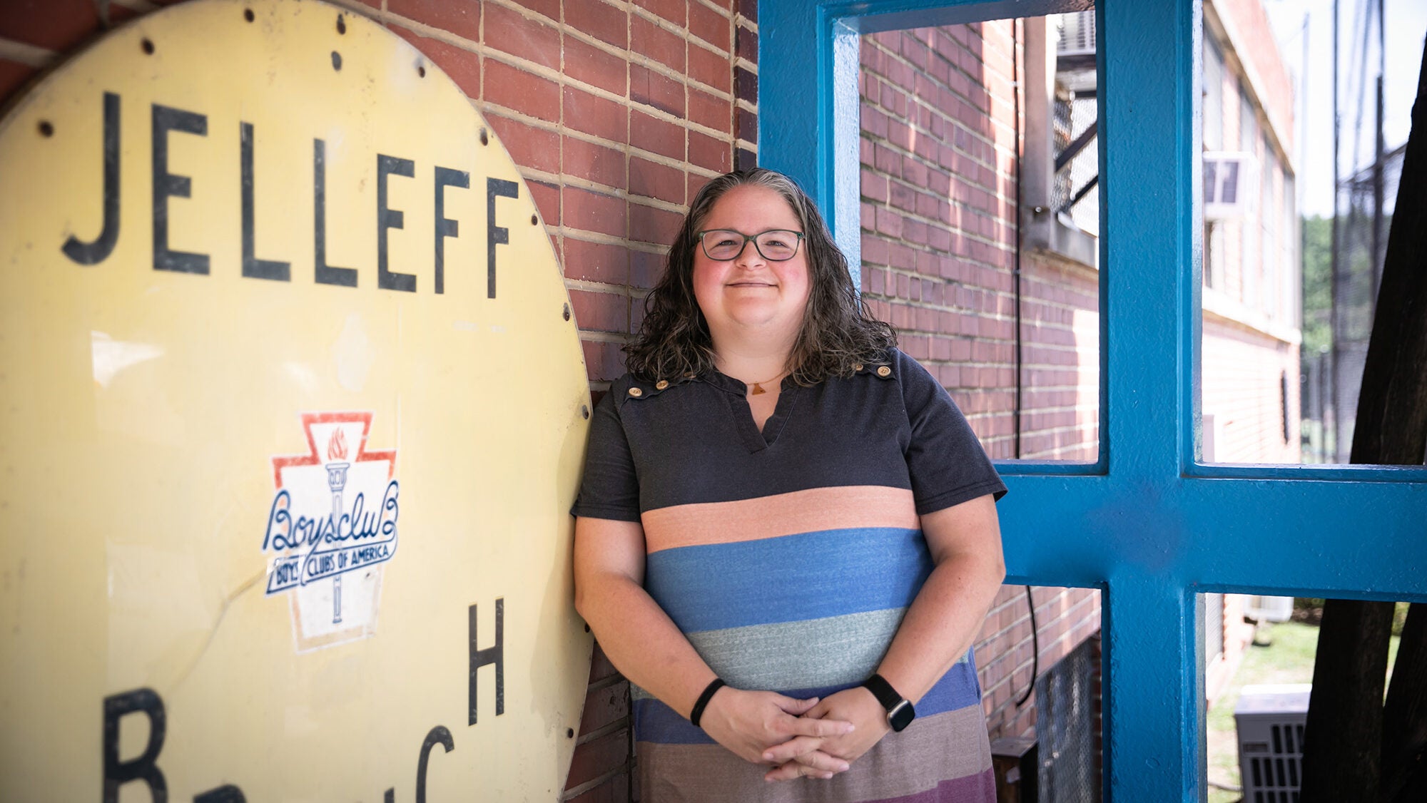 A bespectacled woman with a striped shirt stands in front of a brick wall.