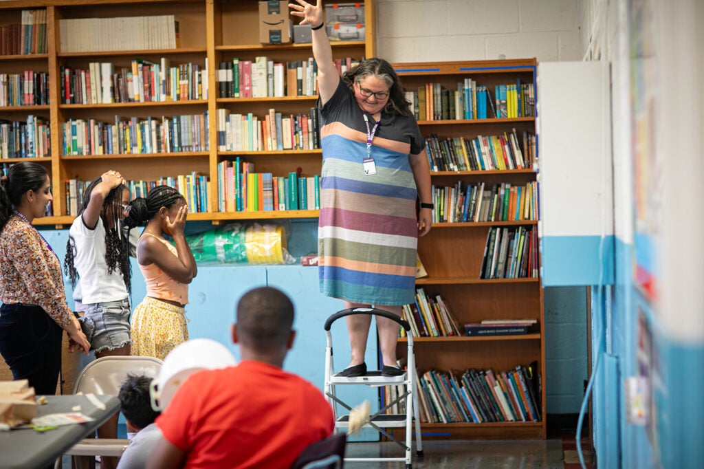 A woman in a striped dress stands in front of book shelves and drops an egg from a step ladder in front of a room full of students. 