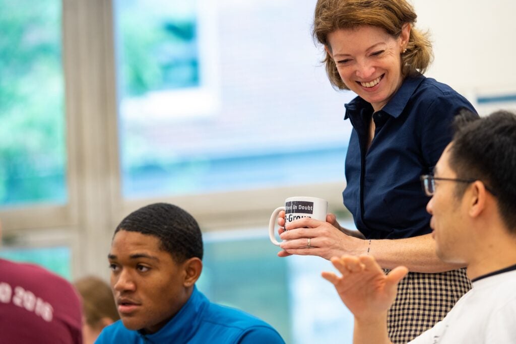 A woman with medium-length hair holds a coffee mug and chats with two male students.