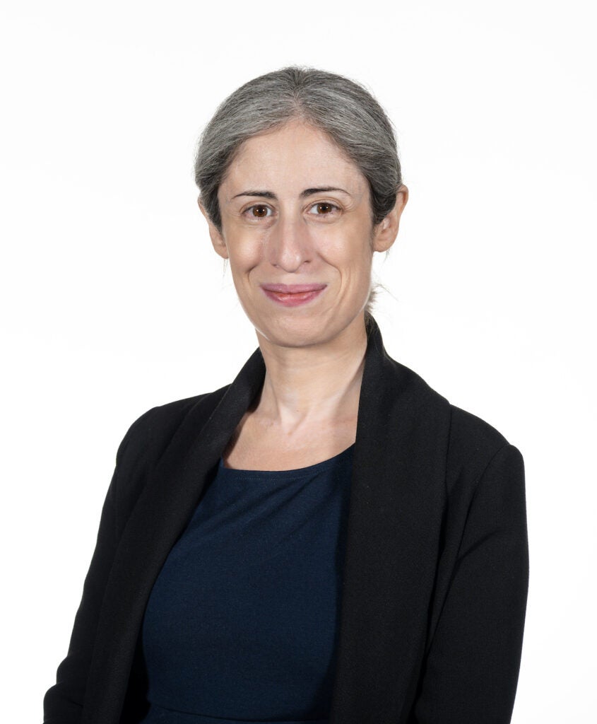 A woman smiles at the camera in front of a white background. She wears smart business attire.