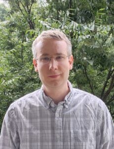 A bespectacled man with short, well-groomed hair standing outside in front of greenery.