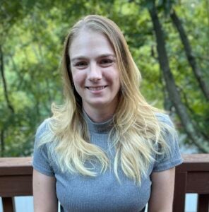 A woman with long, blonde hair stands outside. She wears a gray top. 