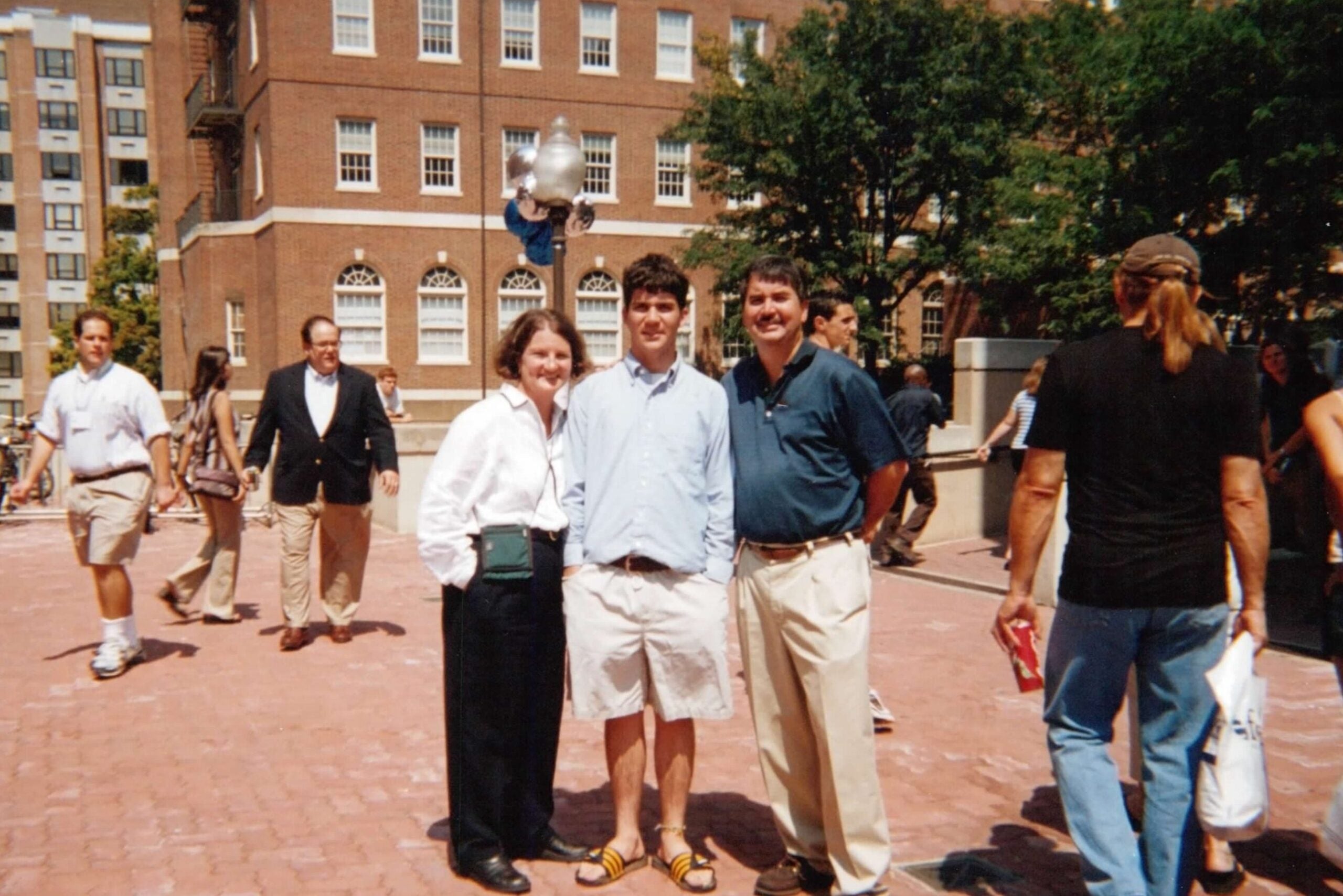 A young man wearing a button down shirt, khaki shorts, and slides stands in between his parents.