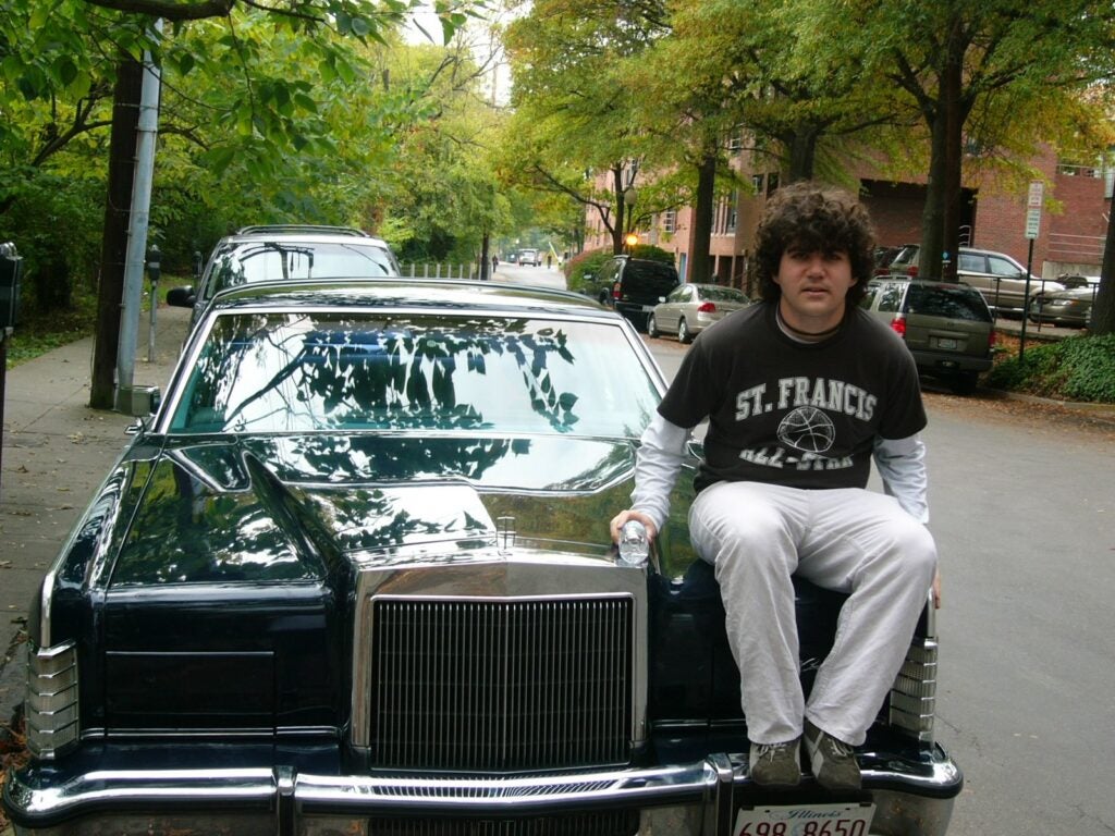 A young man with curly hair sits on the hood of a Buick.  