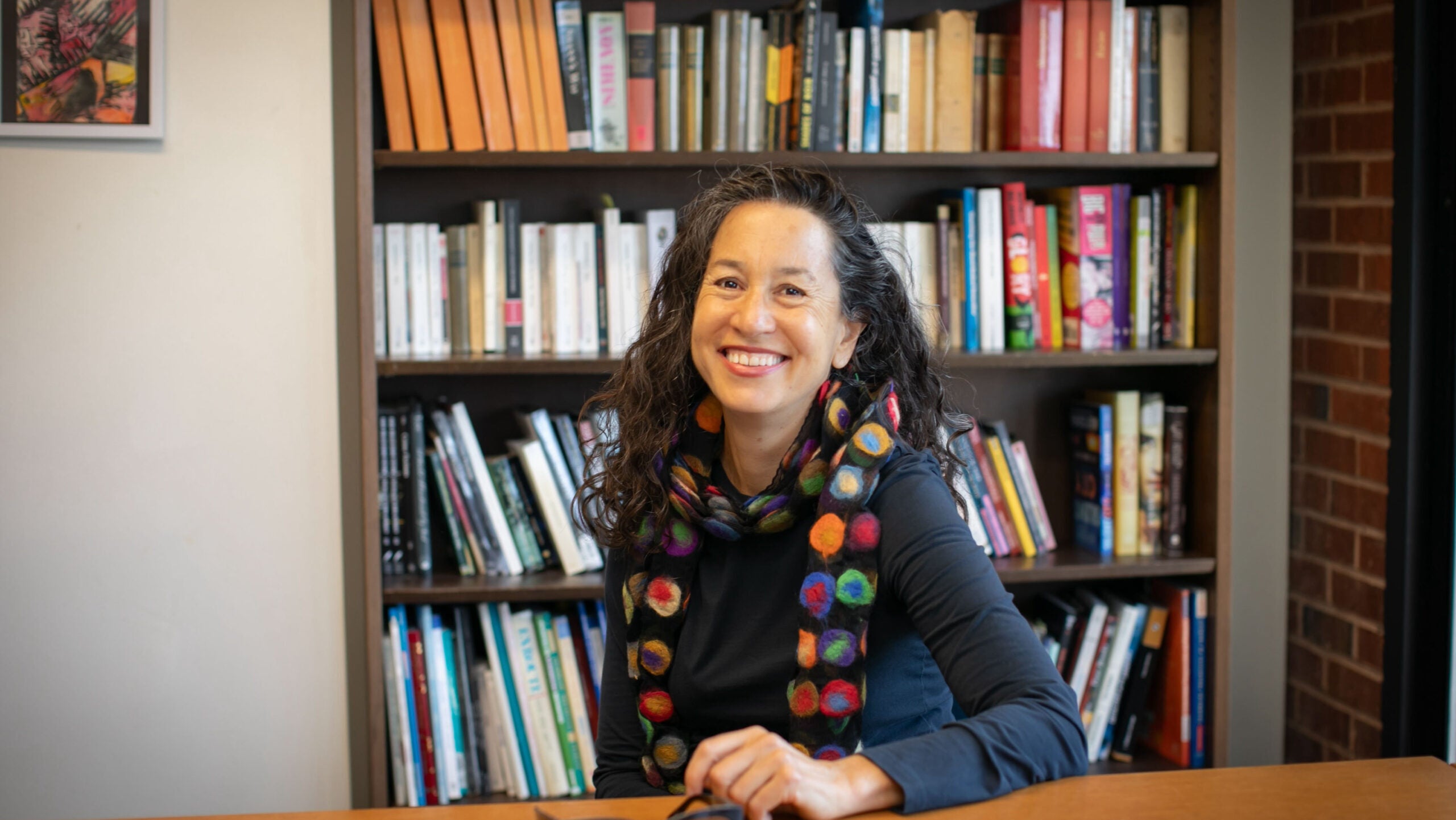 A woman with long, black, curly hair sits at her desk and smiles.