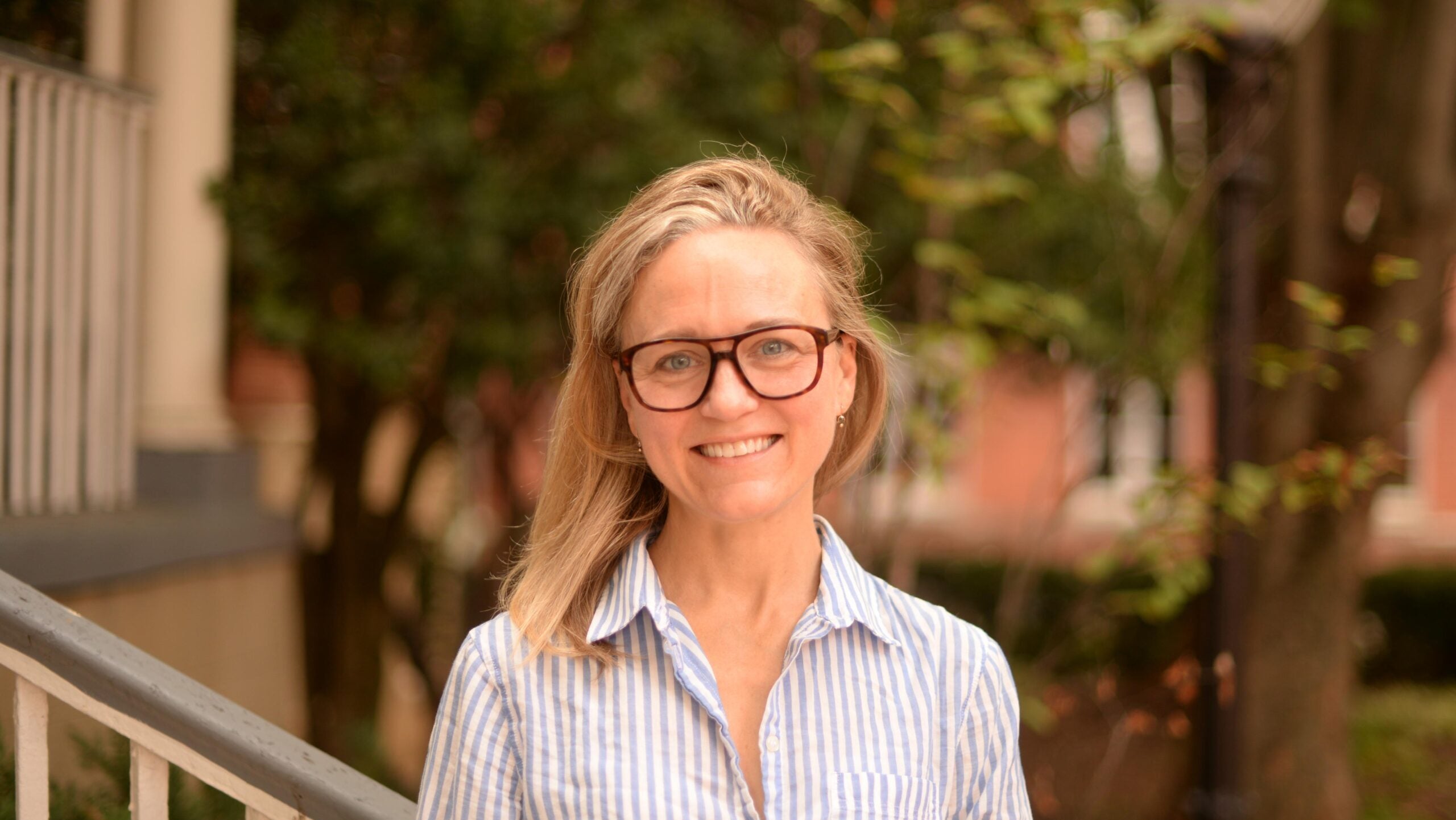 A woman with long, blonde hair smiles. She stands outside and wears a stripped button down shirt that is blue and white.