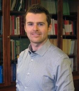 A man with short hair smiles inside in front of a bookshelf. He wears a light blue button down shirt.