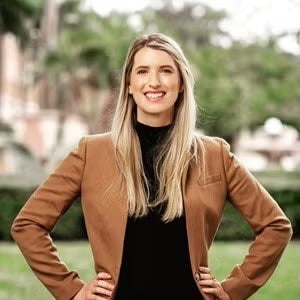 A woman with long blonde hair smiles at the camera. She wears a black top and a tan leather jacket.