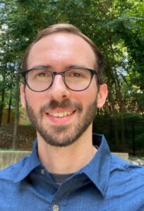 A bespectacled man with short hair stands outside. He wears a blue button down shirt.