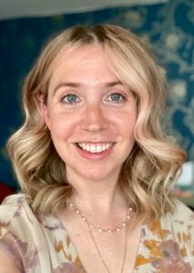 A woman with medium-length blonde hair smiles inside. She wears a blouse with a floral pattern.