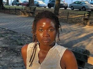 A woman with braids and a white top sits outside and looks at the camera. 