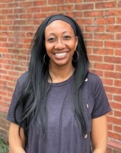 A woman with long, dark hair smiles in front of a red brick wall.