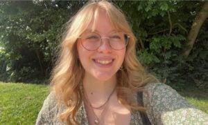 A bespectacled woman with long blonde hair smiles outside. She wears a floral blouse. 