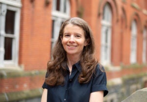 A woman with medium-length hair smiles in front of an out-of-focus red brick wall.