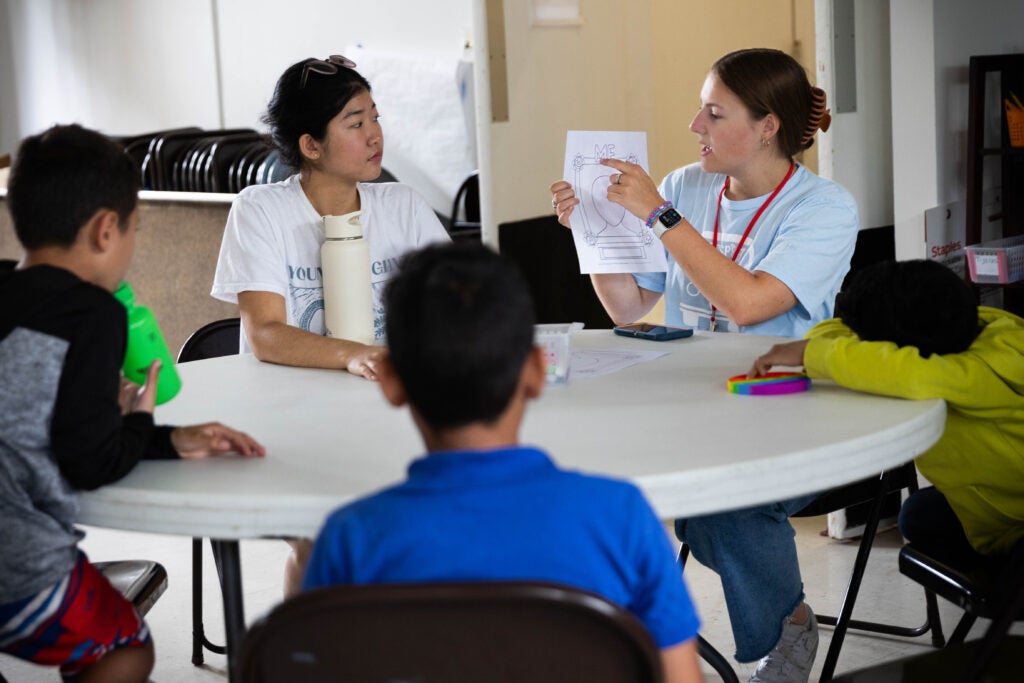 Two college-aged girls sit at a round table with three elementary-school-aged students. Together they are reviewing a worksheet. 