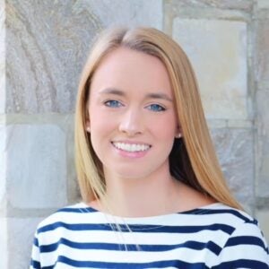 A girl with long blonde hair and blue eyes smiles at the camera. She wears a blue and white striped shirt.