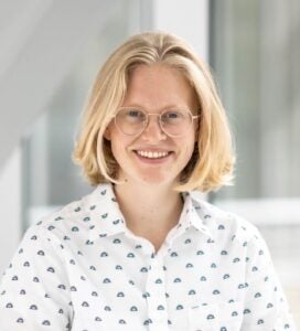 A person with medium-length blonde hair wears a white shirt and smiles at the camera.