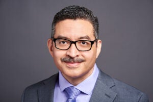 A bespectacled man with short hair softly smiling inside. He wears a jacket and tie. 