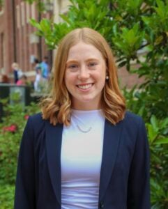 A woman with medium-length red hair smiles outside in front of greenery. She wears a white top and a navy blue blazer.