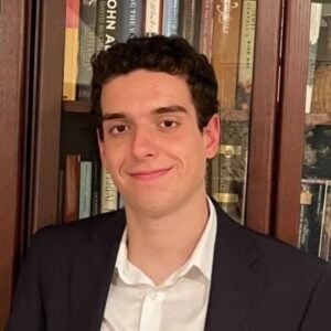 A young man smiles in front of a book case. He wears a white button down and a black suit jacket. 