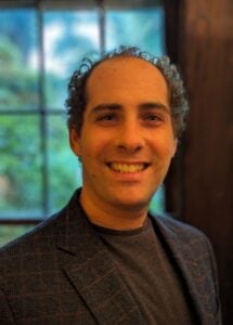 A man with short hair smiles inside. he wears a brown tee shirt and matching blazer. 