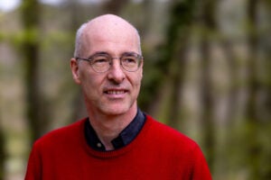 A bespectacled man in a red sweater stands outside and looks away from the camera.