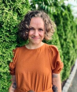 A woman with short, curly hair smiles in front of greenery. She wears an orange romper. 