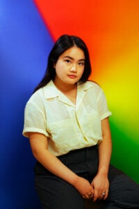 A woman in a white blouse and black pants looks at the camera. She stands in front of a rainbow colored background.