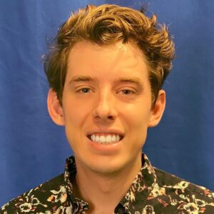 A man with short hair smiles in front of a blue background. He wears a shirt with a floral pattern.