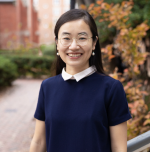 A bespectacled woman with long, dark hair smiles outside. 