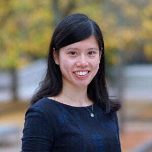 A woman with long dark hair stands outside and smiles. She wears a blue blouse and a silver necklace.